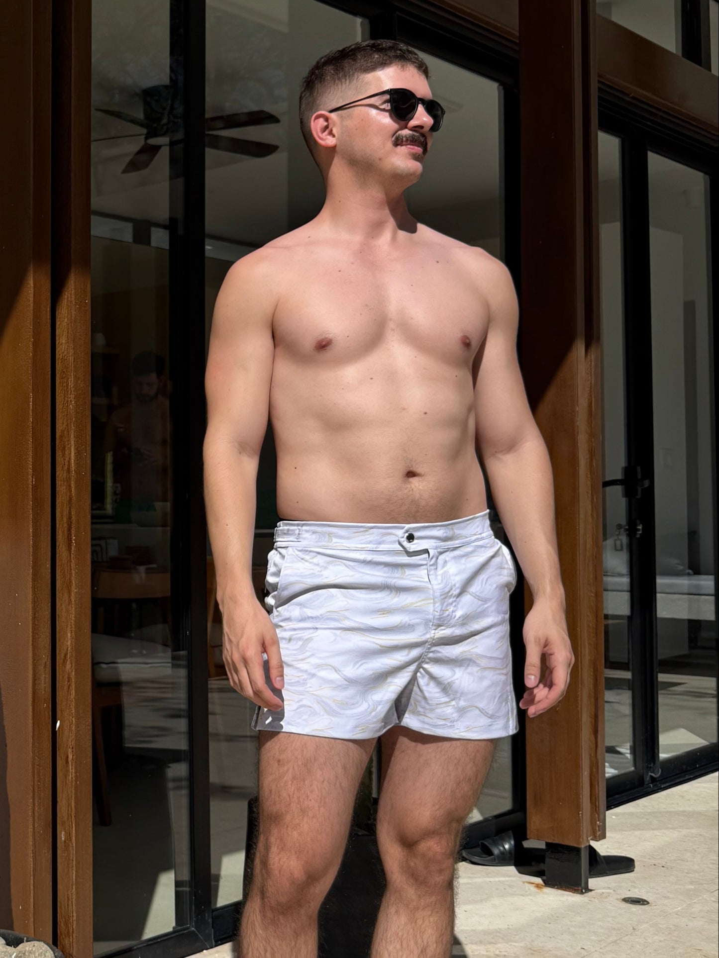 Man standing shirtless in a home setting with a cactus plant in the foreground. wearing swim shorts that are white and gold marble print
