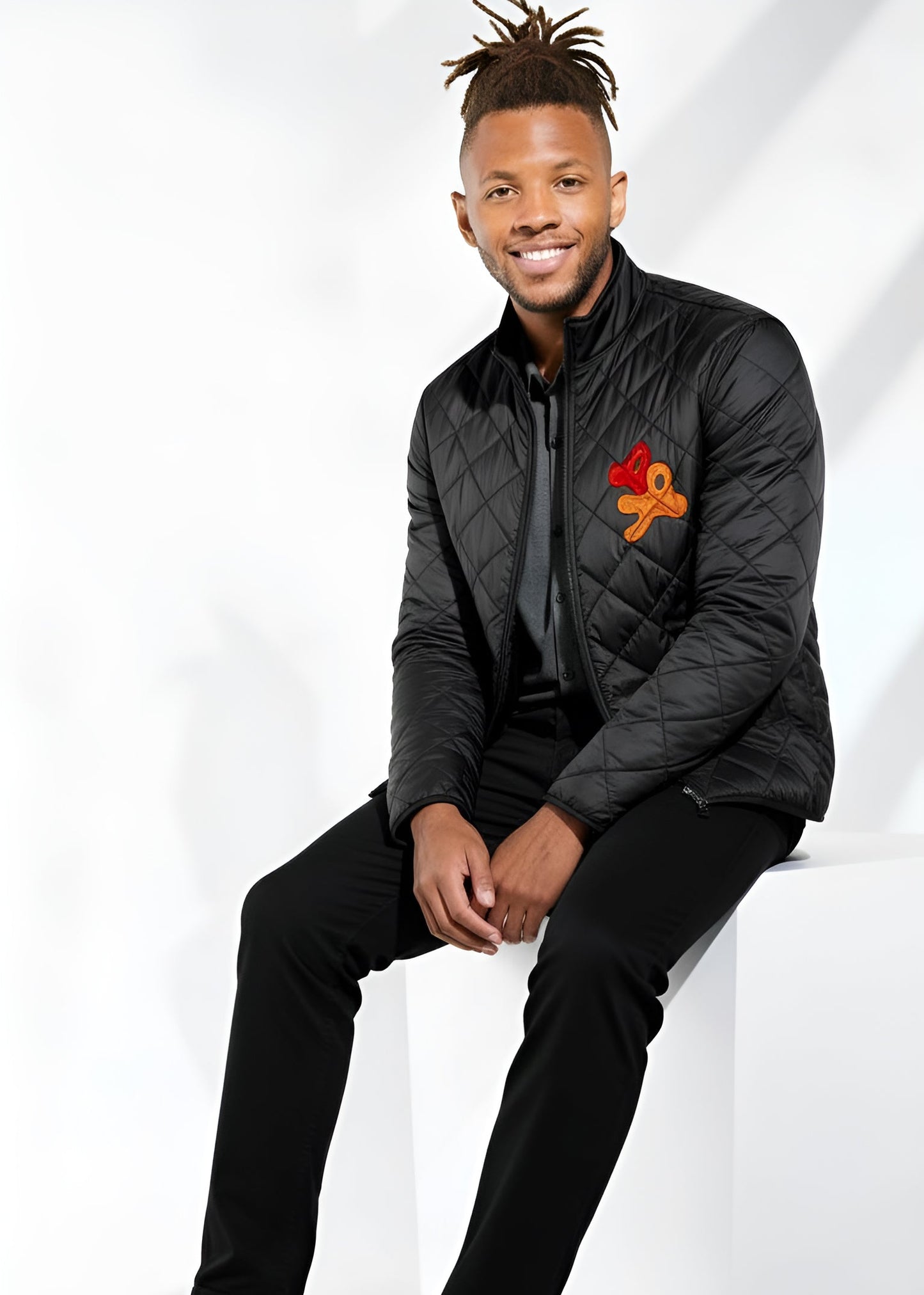 Man wearing a black quilted jacket with a logo, sitting on a white surface.