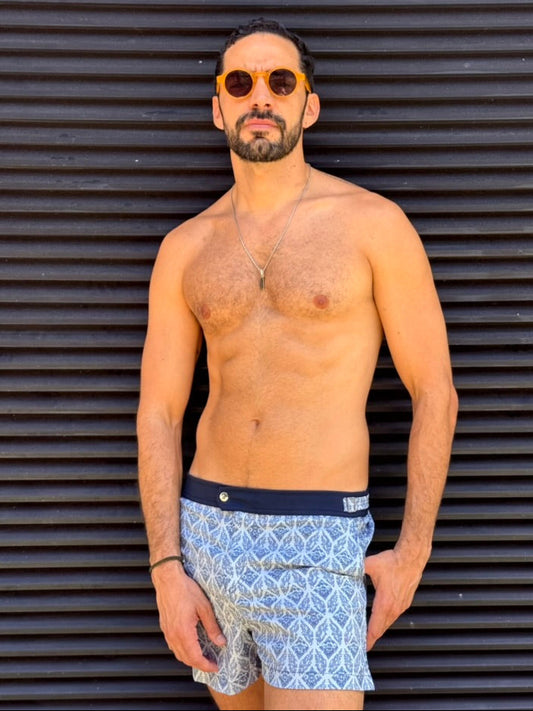 Man wearing sunglasses and patterned swim shorts against a corrugated metal background. luxury men’s swimsuits. Aegean Mosaic pattern