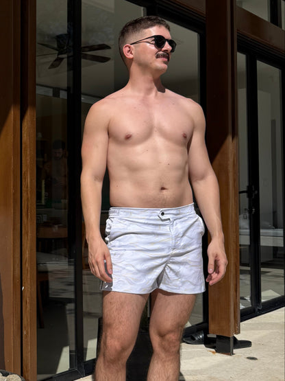 Man standing shirtless in a home setting with a cactus plant in the foreground. wearing swim shorts that are white and gold marble print