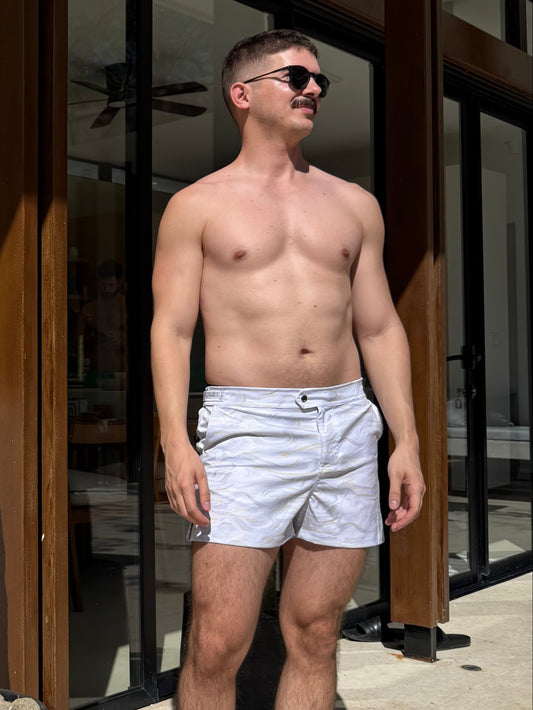 Man standing shirtless in a home setting with a cactus plant in the foreground. wearing swim shorts that are white and gold marble print