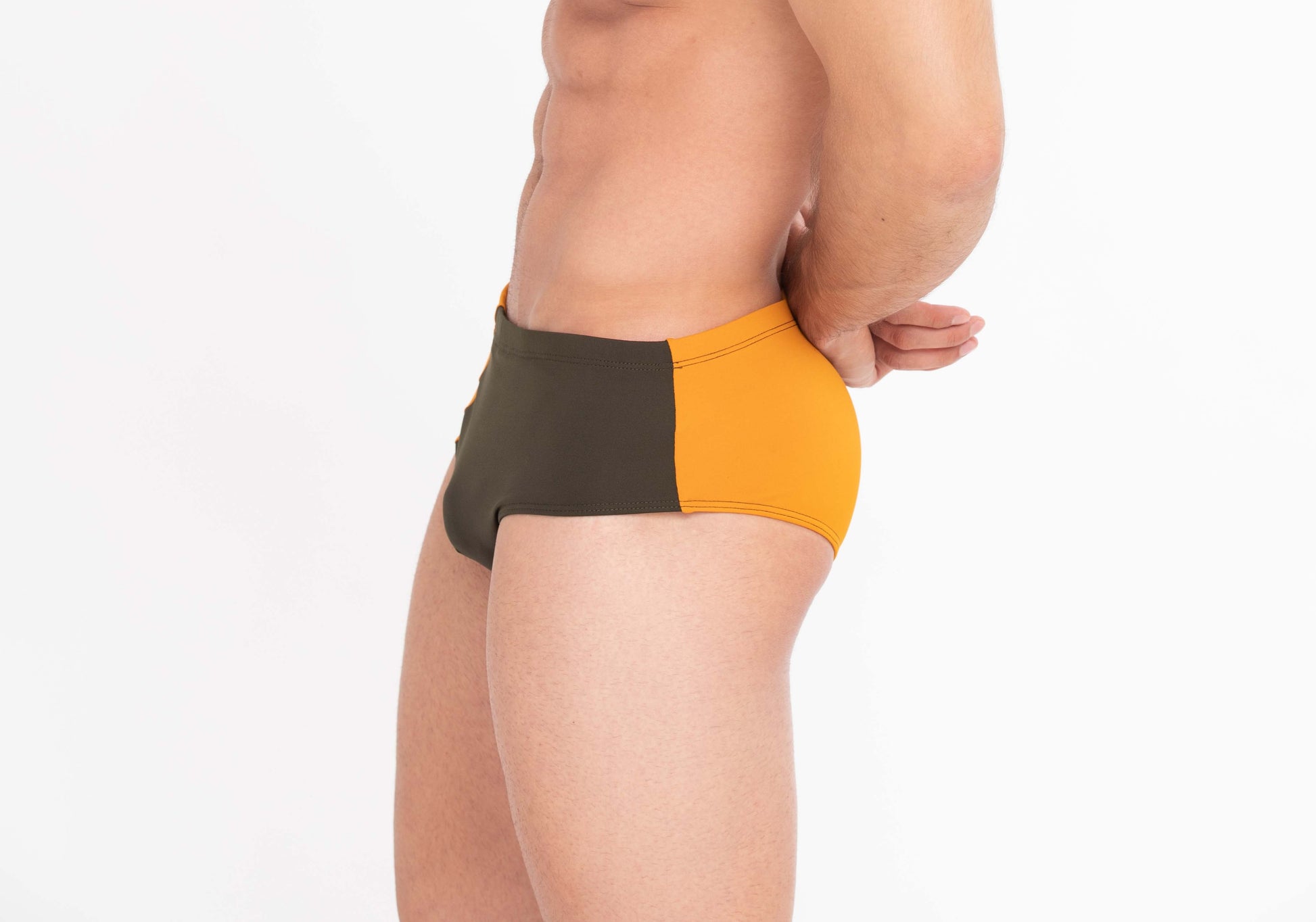 This image features a male model sporting The Athletic Swim Brief, inspired by the vibrant sunsets over Albufeira, Portugal. The swimsuit is designed with a striking color block pattern in sunset orange and olive green, set against a more muted green waistband.